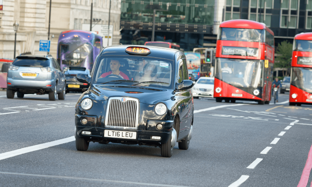 Important things to know London: This is an image of a black cab on a street in London, with cars and red buses behind it.
