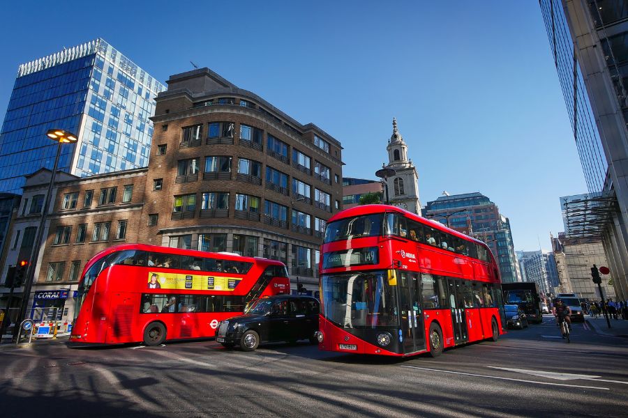 Image of a black cab in between two red London buses, driving on a road in central London. 