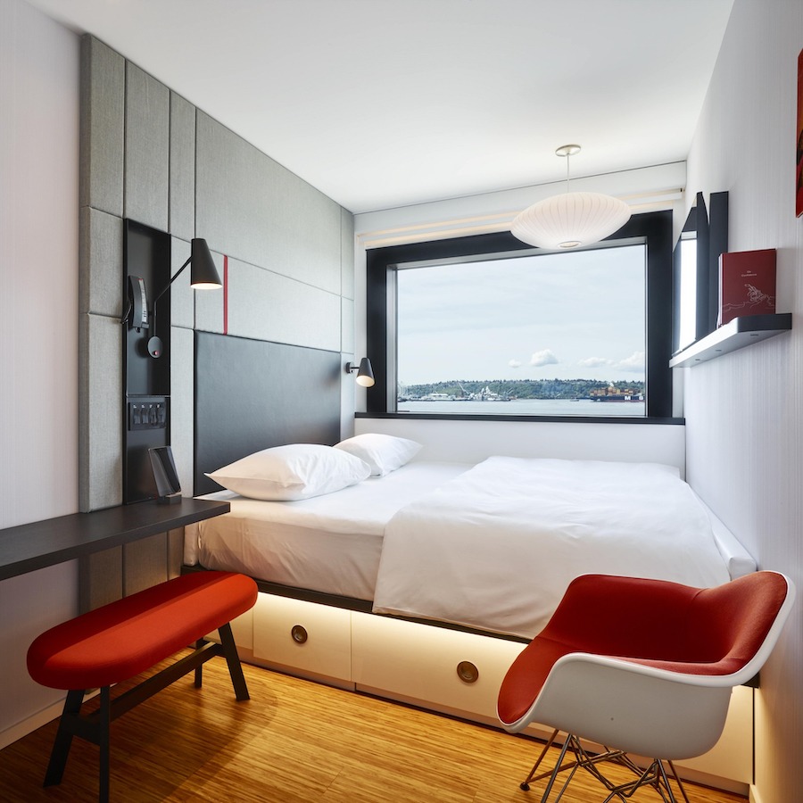 This is an image of a hotel bedroom with a big double bed neatly made and minimalistic furniture toned in hues of red and wooden floors. 