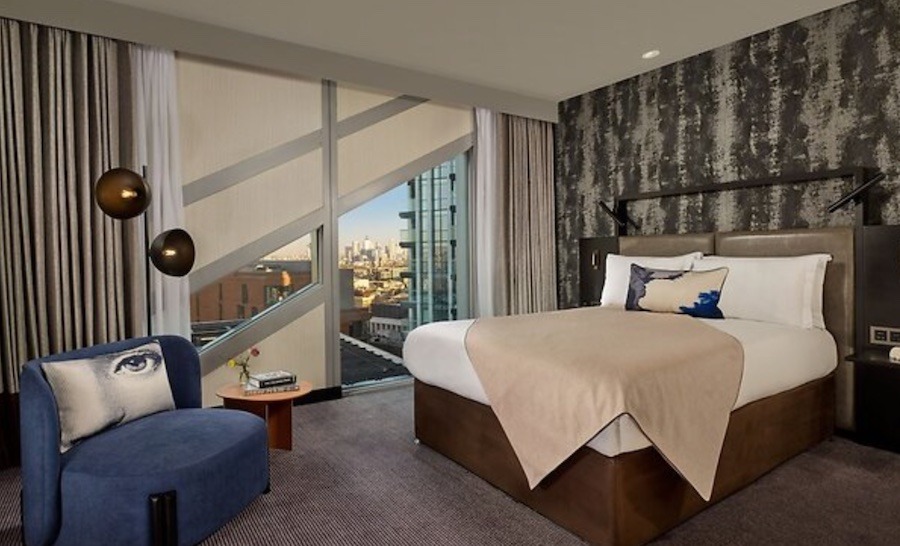 This is an image of a modern hotel room with a double bed that is neatly made, a blue armchair opposite the bed, big glass windows on the right hand side of the bed and a grey carpet covering the floor. 
