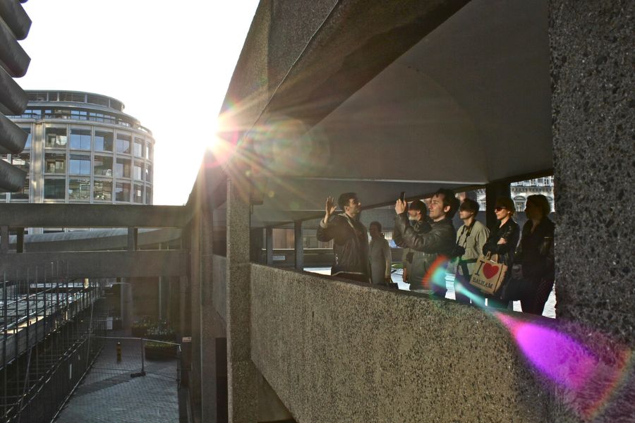 Visitors taking photos from a Barbican Estate walkway in London with sunlight streaming through the brutalist architecture