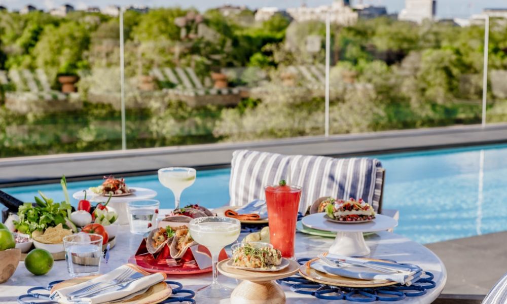 This is an image of a lovely rooftop with a big food and drink spread on the table and a pool in the background.
