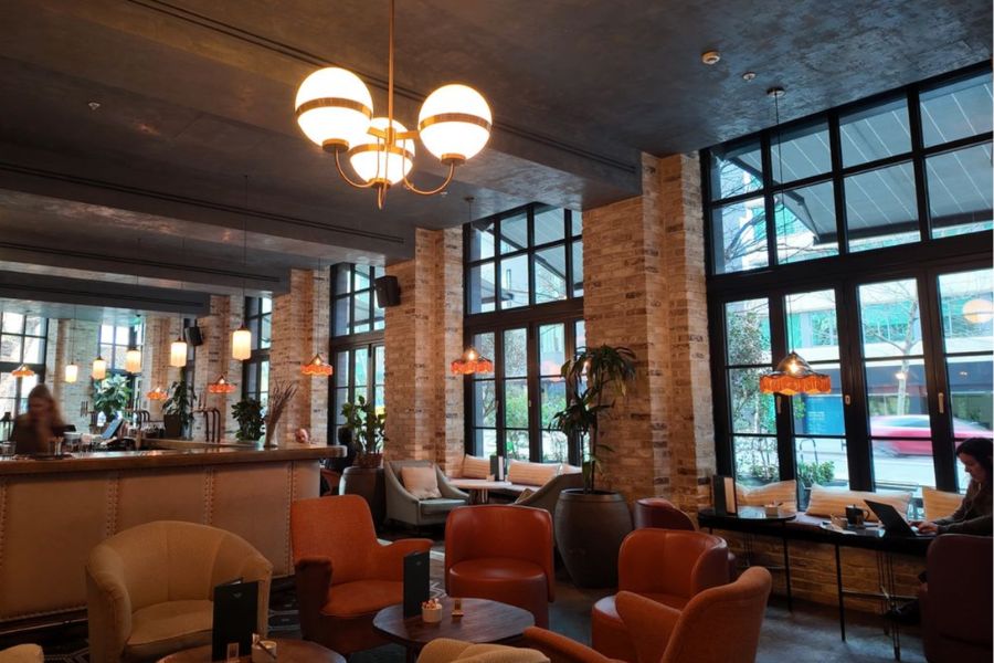 This is an image of a lobby with chairs and tables all around and big industrial windows letting some light in.