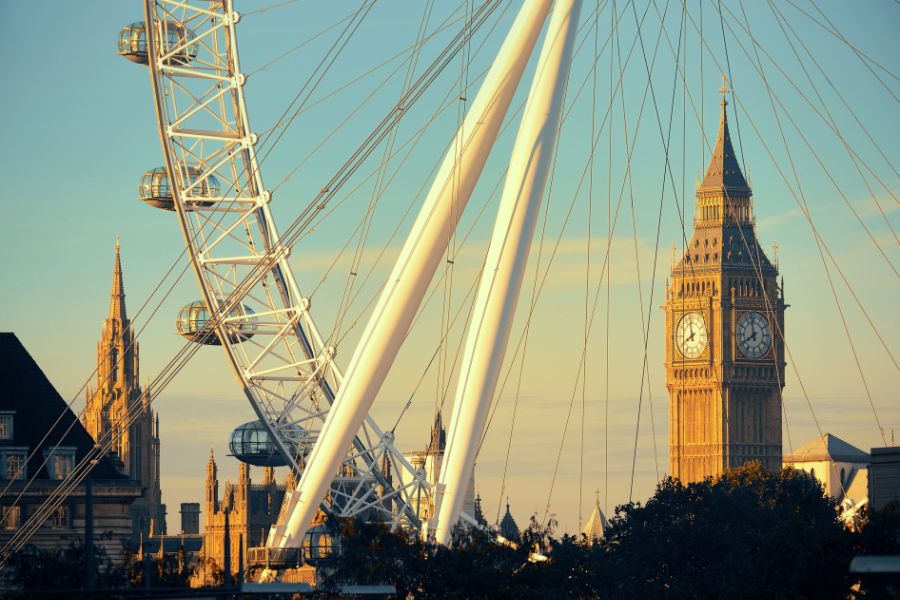 The white steel structure of the London Eye framing Big Ben and the Houses of Parliament bathed in warm golden morning light against a clear blue sky.