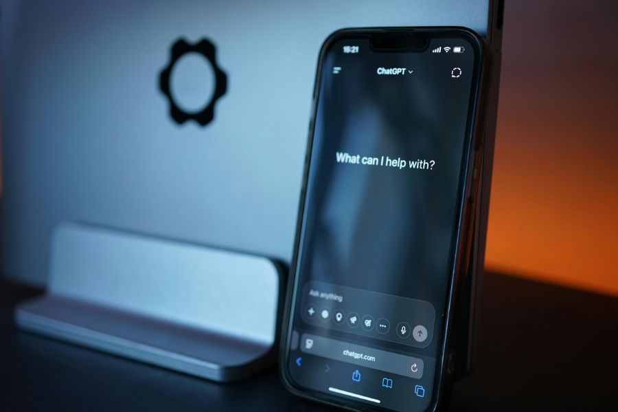 ChatGPT open on a smartphone screen in front of a dark laptop, with the prompt "What can I help with?" displayed