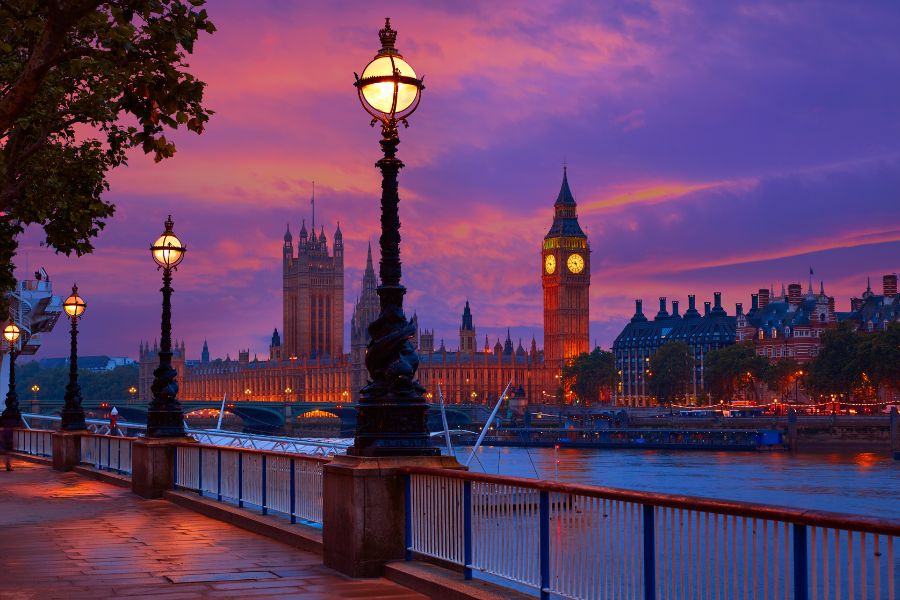 View along the South Bank at dusk, with ornate Victorian lamp posts lining the Thames path and Big Ben and the Houses of Parliament glowing gold against a vivid purple and pink sunset sky.