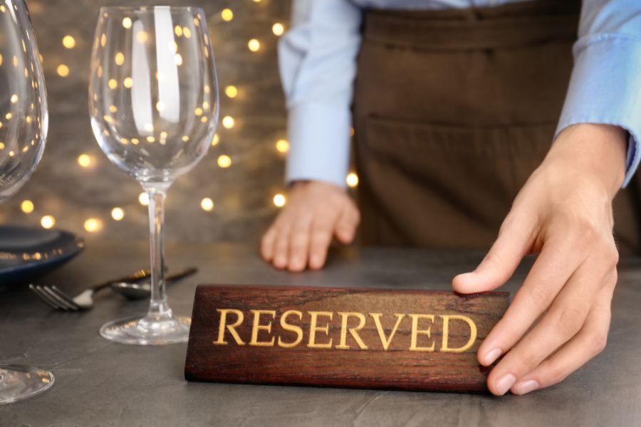 A restaurant server placing a wooden "Reserved" sign on a formally set table with wine glasses and soft fairy lights in the background.
