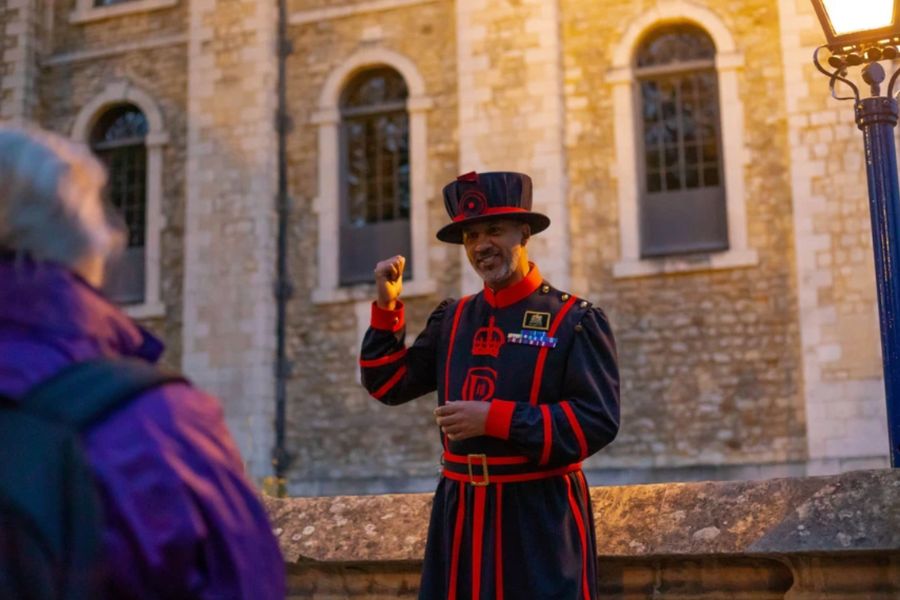 Yeoman Warder (Beefeater) in traditional navy and red uniform gesturing while giving a tour at the Tower of London at dusk