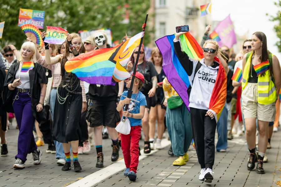 A crowd of people marching in a Pride parade, waving rainbow flags and holding signs including one reading "We're Here We're Queer"