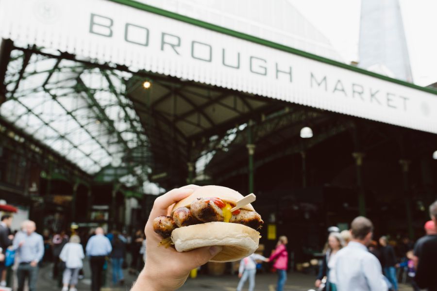 A hand holding a sausage sandwich in front of the Borough Market entrance sign, with crowds of visitors and the market's Victorian iron and glass structure visible behind