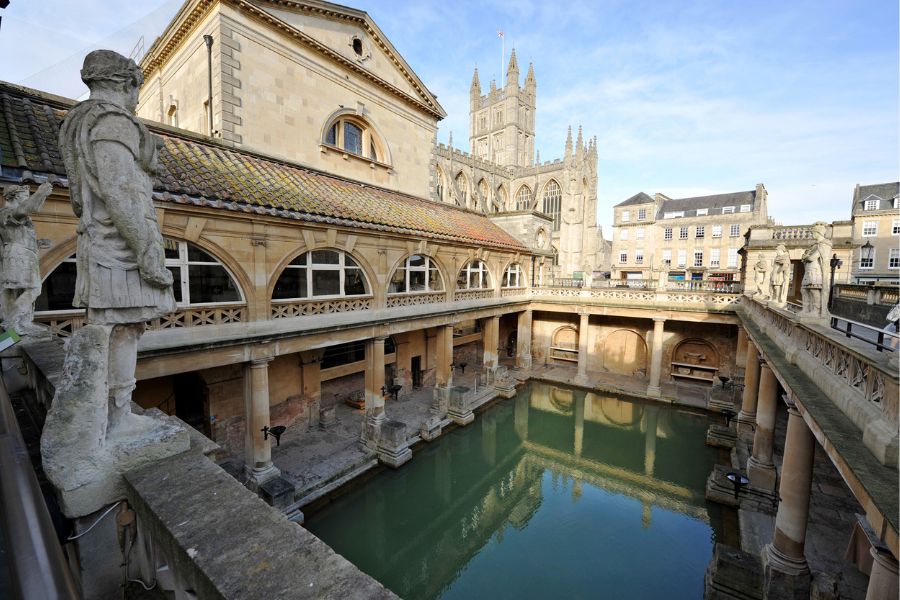 The ancient Roman Baths in Bath, showing the open-air green thermal pool surrounded by classical columns and stone statues, with Bath Abbey visible in the background