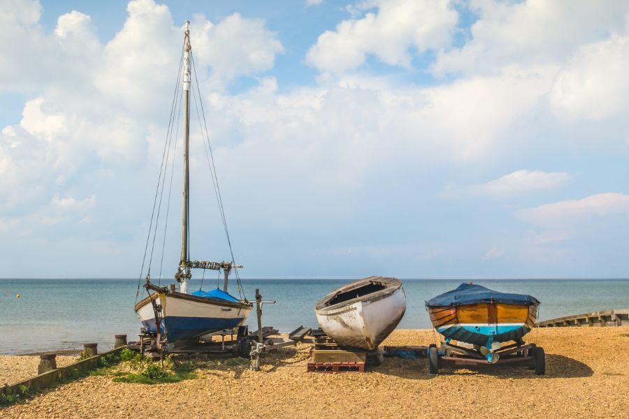 Three small wooden boats resting on a shingle beach in Whitstable on a sunny day, with the calm sea stretching out behind them