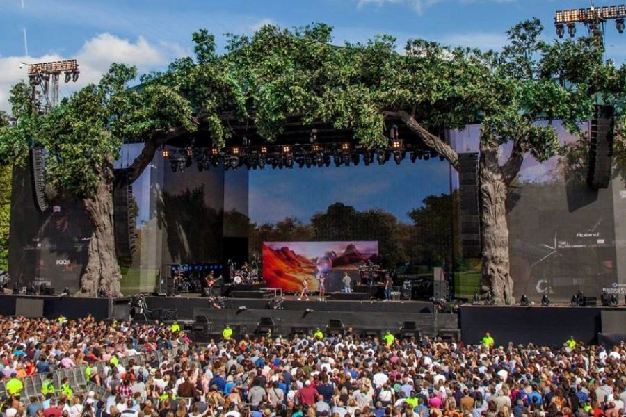 A packed crowd watching a live performance on the BST Hyde Park main stage, which is decorated with large sculpted trees along its sides