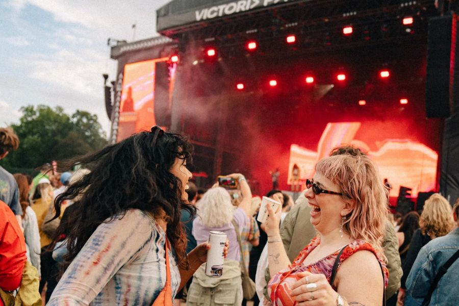 Two women laughing and holding drinks cans in front of a brightly lit stage at All Points East festival in Victoria Park