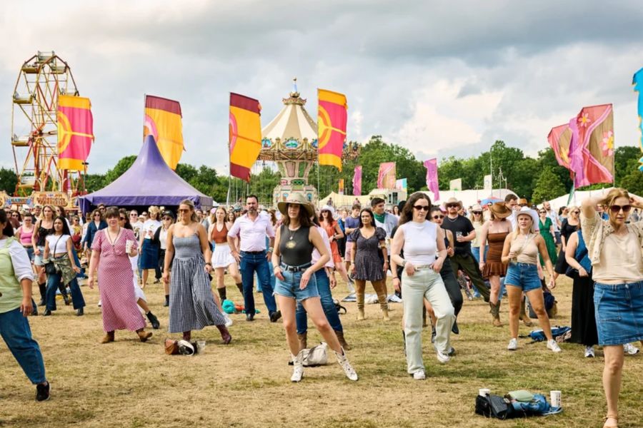 A crowd of festival-goers line dancing on a grass field at Country on the Common, with colourful flags, a ferris wheel and a carousel visible in the background