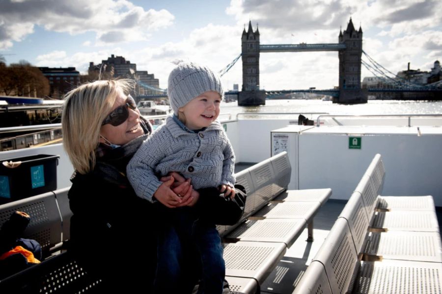 A woman and a young child smiling on the open deck of a Thames river cruise, with Tower Bridge visible in the background
