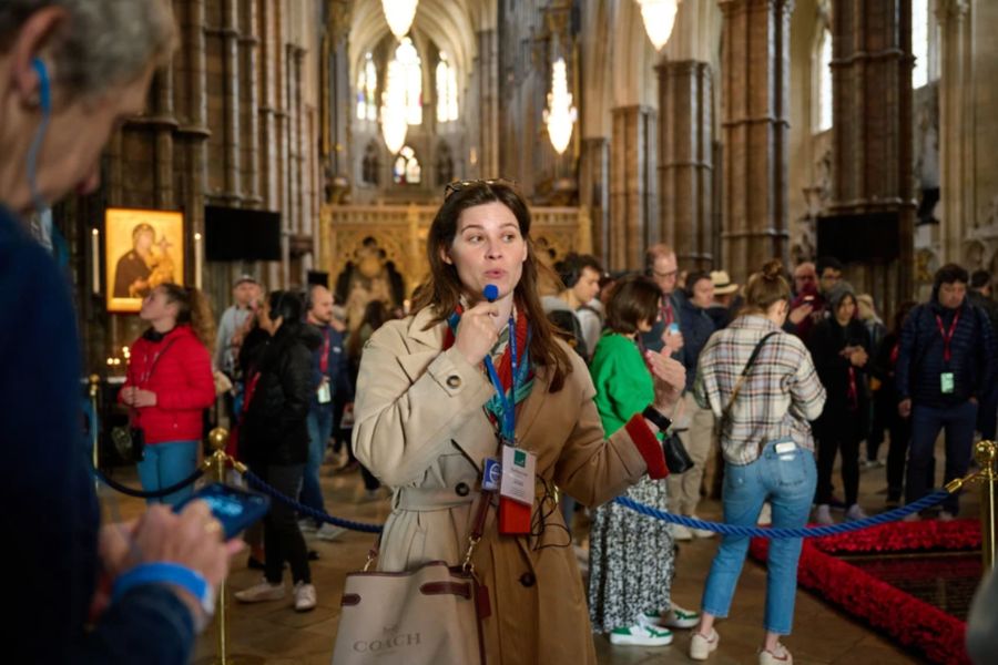 A tour guide speaking into a microphone inside Westminster Abbey, with ornate Gothic architecture and a crowd of visitors visible behind her