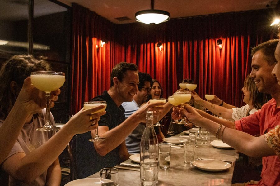 A group of people laughing and clinking cocktail glasses together at a long dinner table in a dimly lit restaurant with red velvet curtains