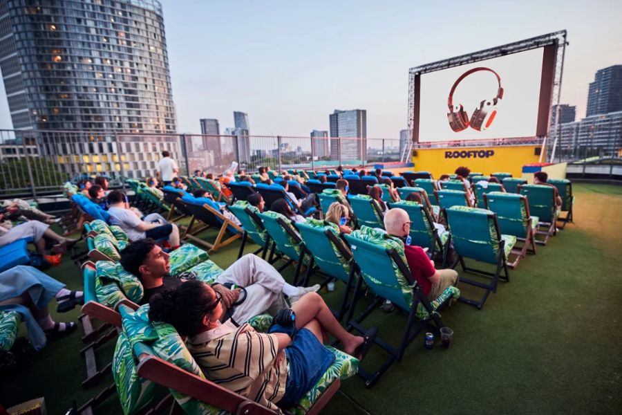 An audience relaxing in tropical-print deck chairs at the Rooftop Cinema Club in London, with the city skyline visible in the background at dusk