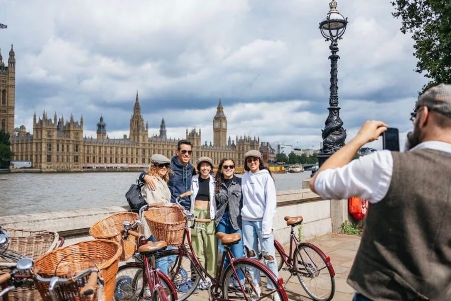 A group of four friends posing with vintage bicycles on the South Bank, with the Houses of Parliament and Big Ben visible across the Thames behind them