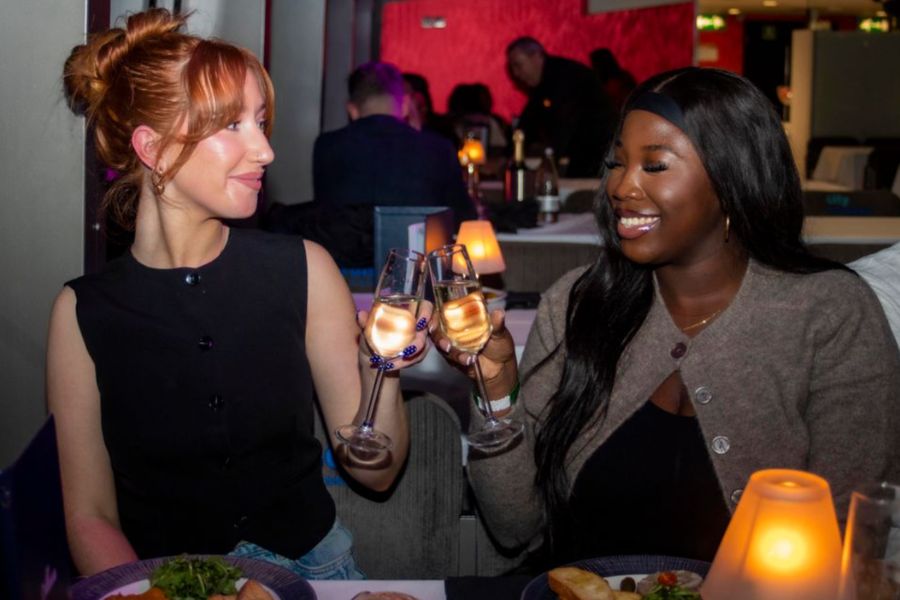 Two women smiling and clinking champagne glasses at a candlelit dinner table on an evening river cruise