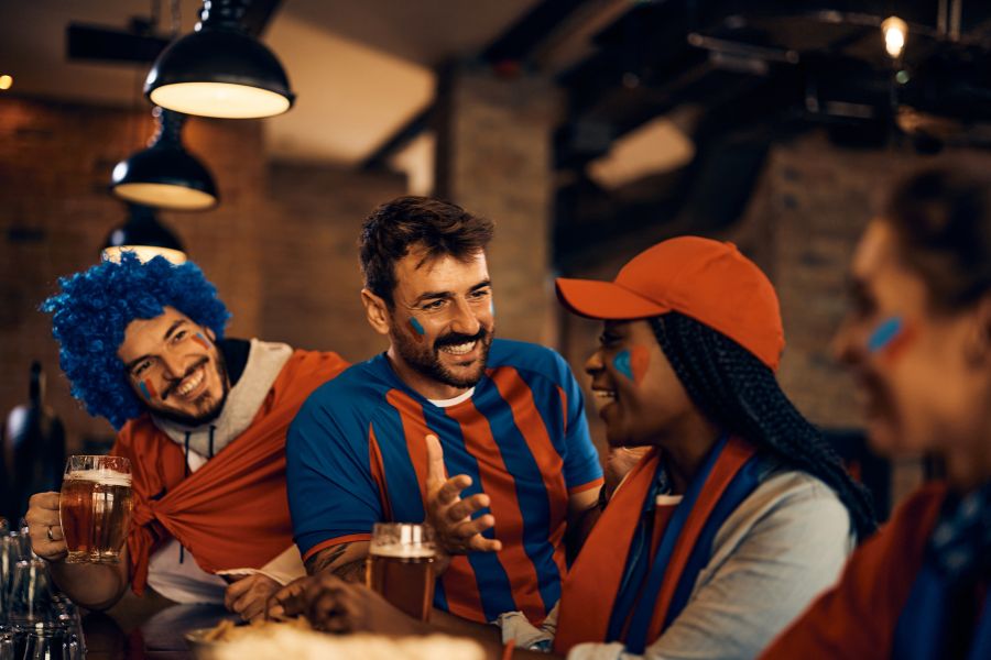 A group of football fans in team colours and face paint laughing together over pints of beer at a pub