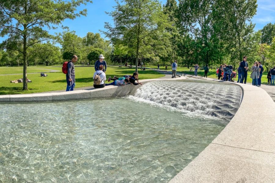 Visitors sitting and playing beside the Diana Memorial Fountain in Hyde Park on a sunny day, with water cascading over the curved granite channel