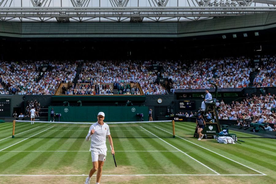 A tennis player in all-white standing on Centre Court at Wimbledon, with a packed crowd of spectators filling the stands behind them