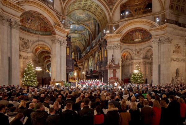 This is an image of a big cathedral hall with people filling all the seats. There are high ceilings full of beautifully carved architecture and a big Christmas tree.