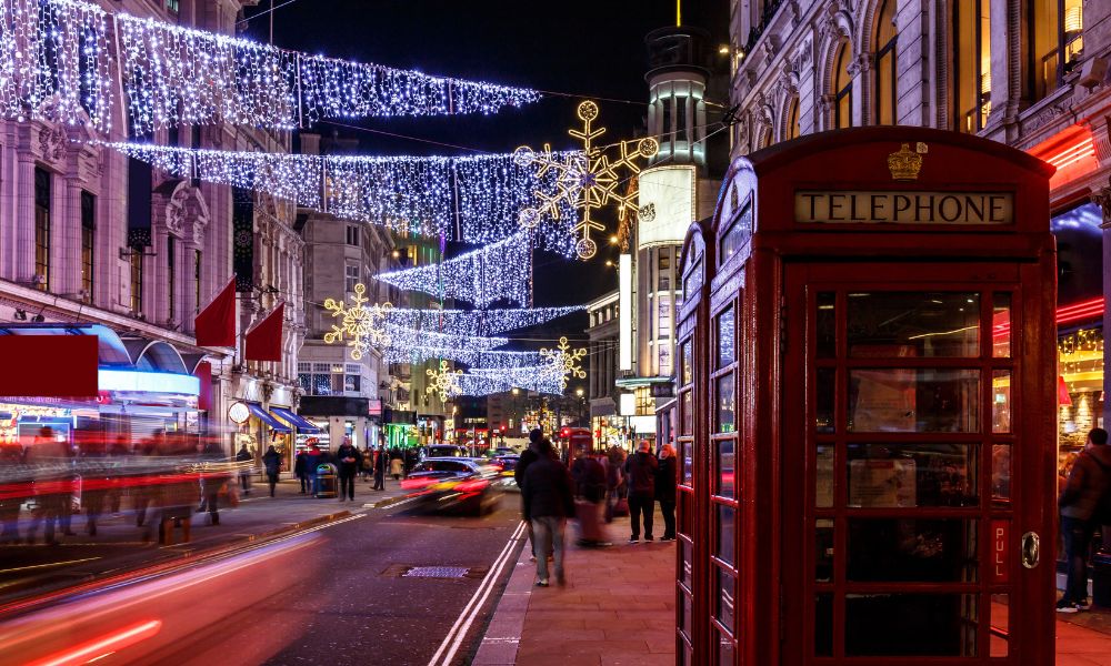 This is an image of the beautiful hanging lights on Oxford Street. There's people walking in the street behind it. There's also a red phone box and a big red double decker bus.