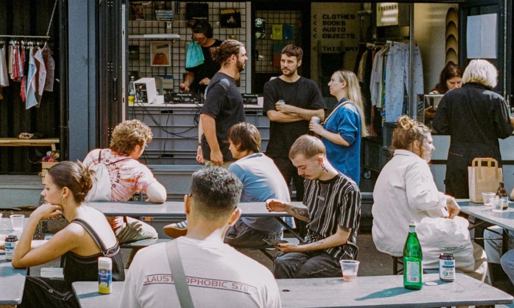 This is an image of a bunch of people mingling outside on wooden benches with food.
