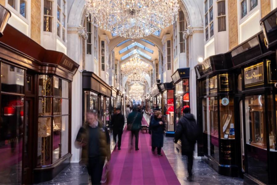 Interior of Burlington Arcade in London, a covered shopping gallery with a pink carpet runner, ornate white arched ceiling adorned with cascading crystal chandeliers, and elegant boutique shopfronts on either side, with shoppers walking through.