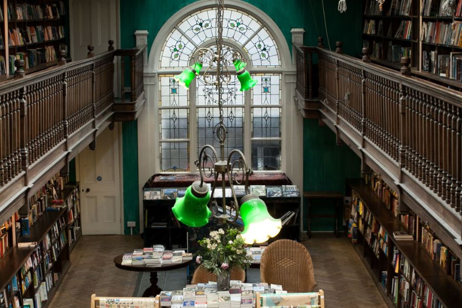 Interior of a grand two-storey independent bookshop with dark green walls, wooden gallery balconies lined with bookshelves, a large arched stained-glass window, and a decorative green glass chandelier hanging above a central table covered in books.