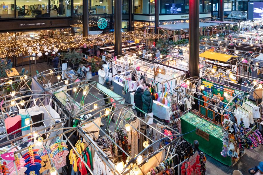 Aerial view of a bustling indoor market hall filled with colourful stalls selling clothing and gifts, with warm fairy lights strung overhead and a bar and restaurant visible in the background.