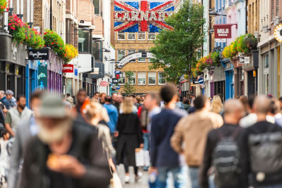 A busy pedestrianised Carnaby Street in London's Soho, with a large Union Jack sign reading "Carnaby" spanning the street and crowds of shoppers walking beneath it, flanked by colourful shopfronts.