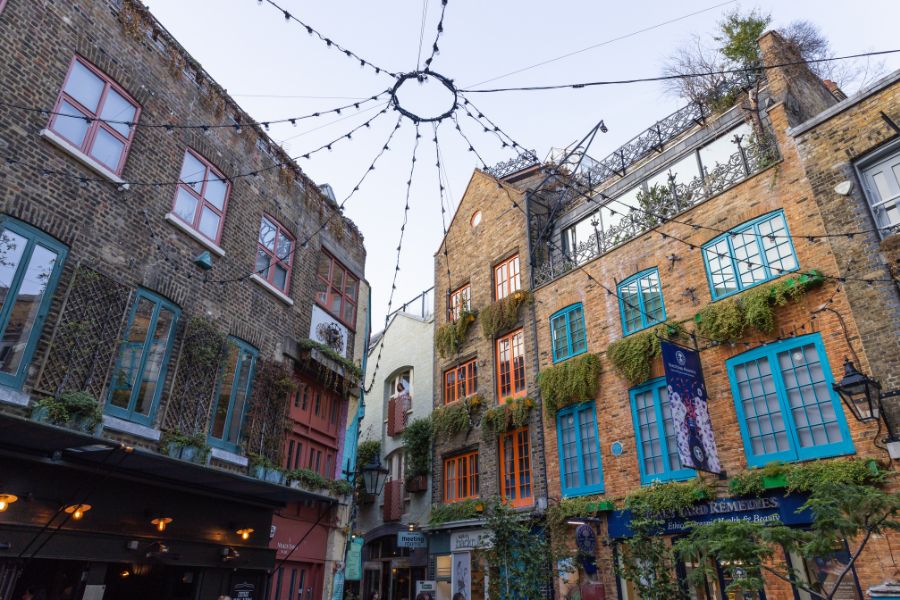 Neal's Yard in Covent Garden, a charming courtyard with tall Victorian brick buildings painted in bright colours, teal and orange window frames draped with climbing plants, and fairy lights strung across the sky above.