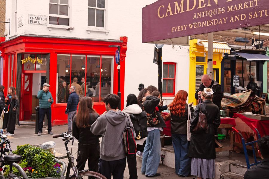 Street scene at Camden Passage Antiques Market in Islington, with shoppers browsing an outdoor stall beneath a burgundy sign advertising the market every Wednesday and Saturday, surrounded by red and yellow shopfronts.