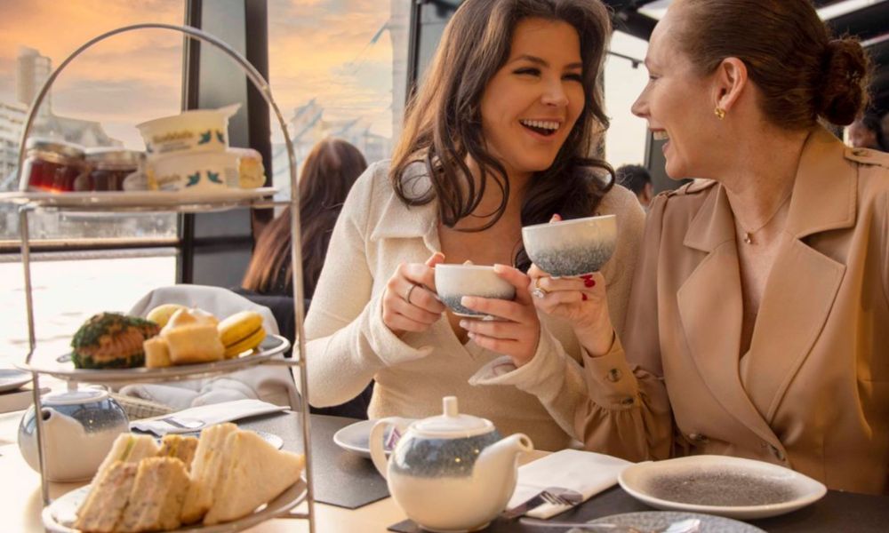 Unique afternoon teas in London: This is an image of a friendship duo laughing together and having afternoon tea on a boat with a stand full of cakes in front of them.