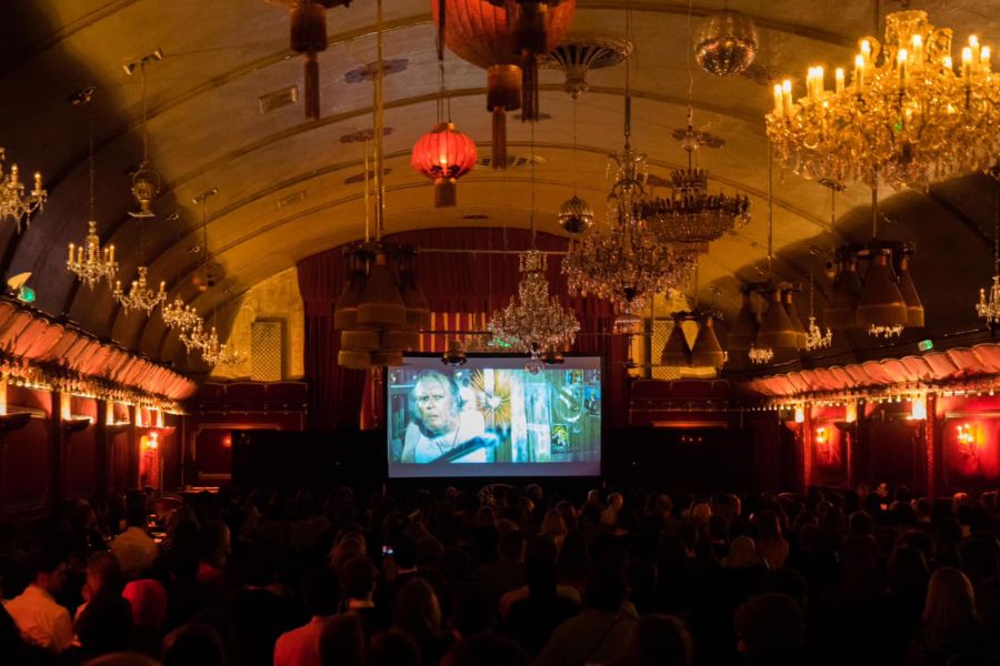 This is an image of an old ballroom cinema with chandeliers and elegant features with a big projector screen at the front.