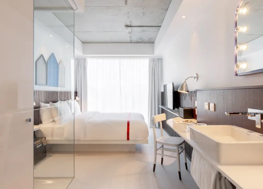 This is an image of a big hotel bedroom with a double bed, walk in shower, white furnishings and big double windows that fill the room with daylight.