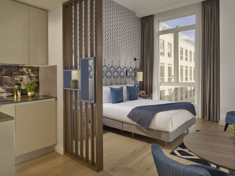 Best hotels in London for long stays: This is an image of a double bed close to a big window with light coming through. There is a dining table to the side, and a small kitchenette with a wooden divider.