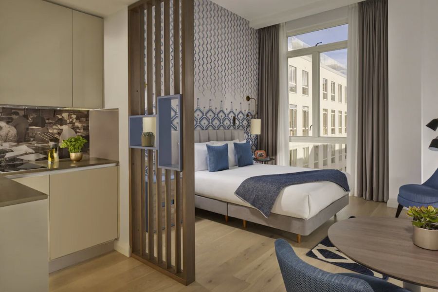 Best hotels in London for long stays: This is an image of a double bed close to a big window with light coming through. There is a dining table to the side, and a small kitchenette with a wooden divider.