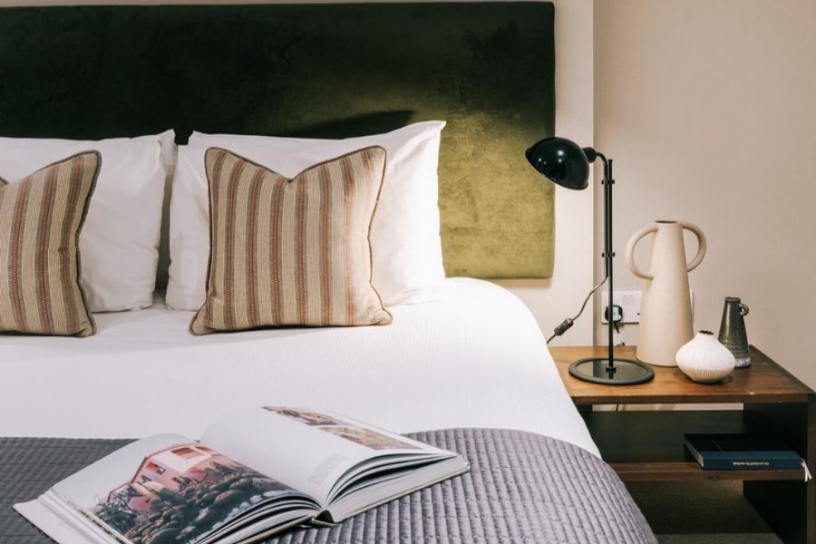 This is an image of a double bed with white sheets, striped cushions and a magazine laying atop of it. There is also a bedside table with a lamp.
