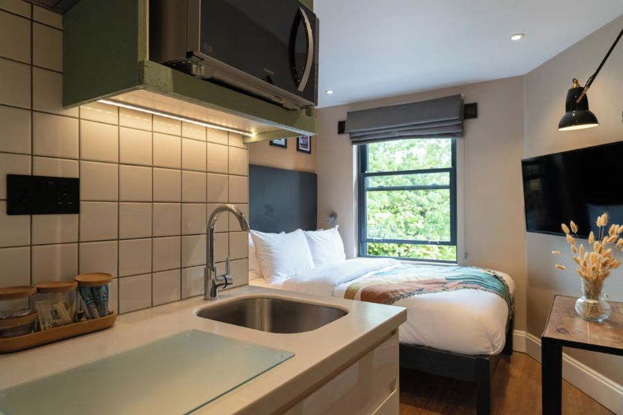 Best hotels in London for long stays: This is an image of a small apart-hotel room with a bed pushed up by the window, with light filtering through. Closer to the entrance, there is a small kitchenette with a sink and chopping board.