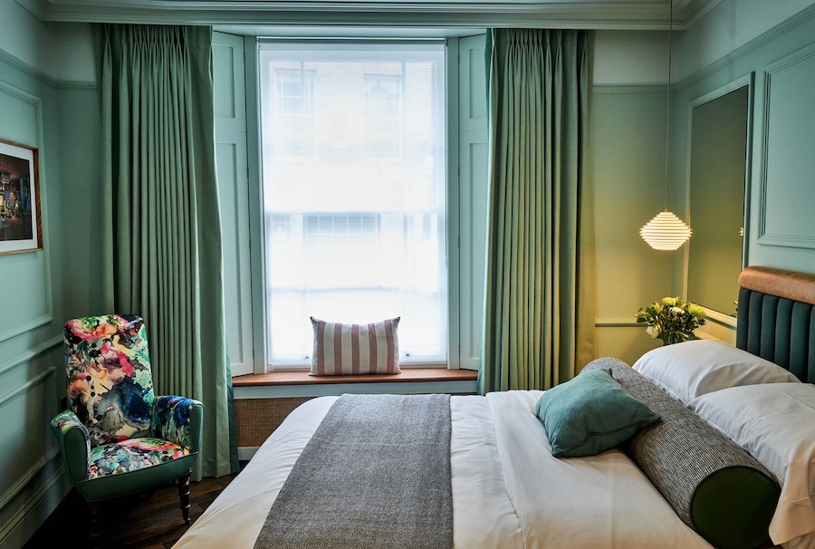 This is an image of a hotel bedroom with a big double bed. The walls and curtains are green as well as the armchair in the corner of the room. It is cool and stylish and unique looking.