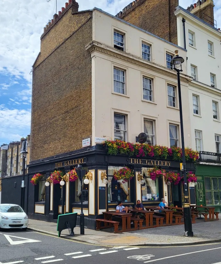 Picture of the exterior of The Gallery, one of the best London hostels, with brick walls and white facade. There’s a pub on the ground floor with flowers all around.