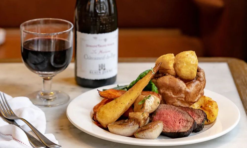 This is an image of a nice roast beef with potatoes and vegetables. There is also an expensive bottle of wine in the background.