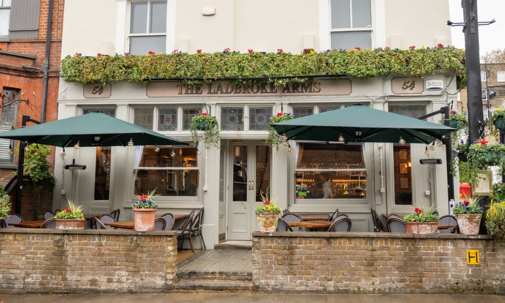 This is an image of a pub with a front porch that has umbrellas over the top of outdoor seating.