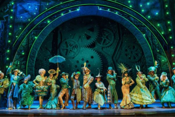 Best West End shows in London: This is an image of the Wicked cast dancing in the middle of the stage with green costumes and lighting, with cogs on the screen behind them.