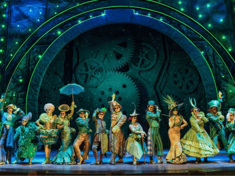 Best West End shows in London: This is an image of the Wicked cast dancing in the middle of the stage with green costumes and lighting, with cogs on the screen behind them.